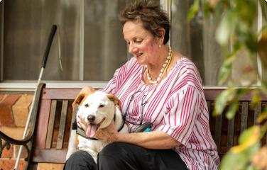 A smiling older woman sits on a bench, petting a white and brown dog. A white cane leans beside her. 