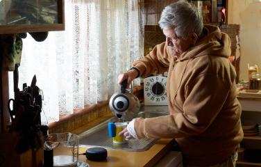 An older man in a brown hoodie pours hot water from a kettle into a cup at the kitchen sink. A talking clock and a smart speaker are nearby.