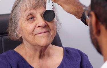 An older woman with grey hair and a warm smile undergoes an eye examination. A healthcare professional holds an occluder over one of her eyes.