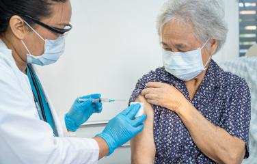 An older woman receiving a vaccination in her upper arm, administered by a doctor wearing gloves. Both are wearing masks.