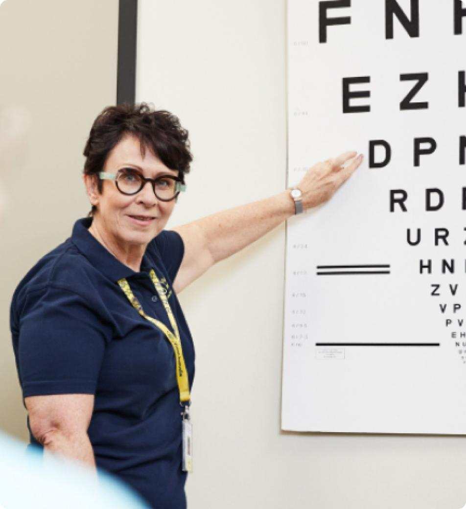 A woman in a navy polo shirt points to an eye chart on the wall. She wears glasses and a lanyard, smiling slightly.