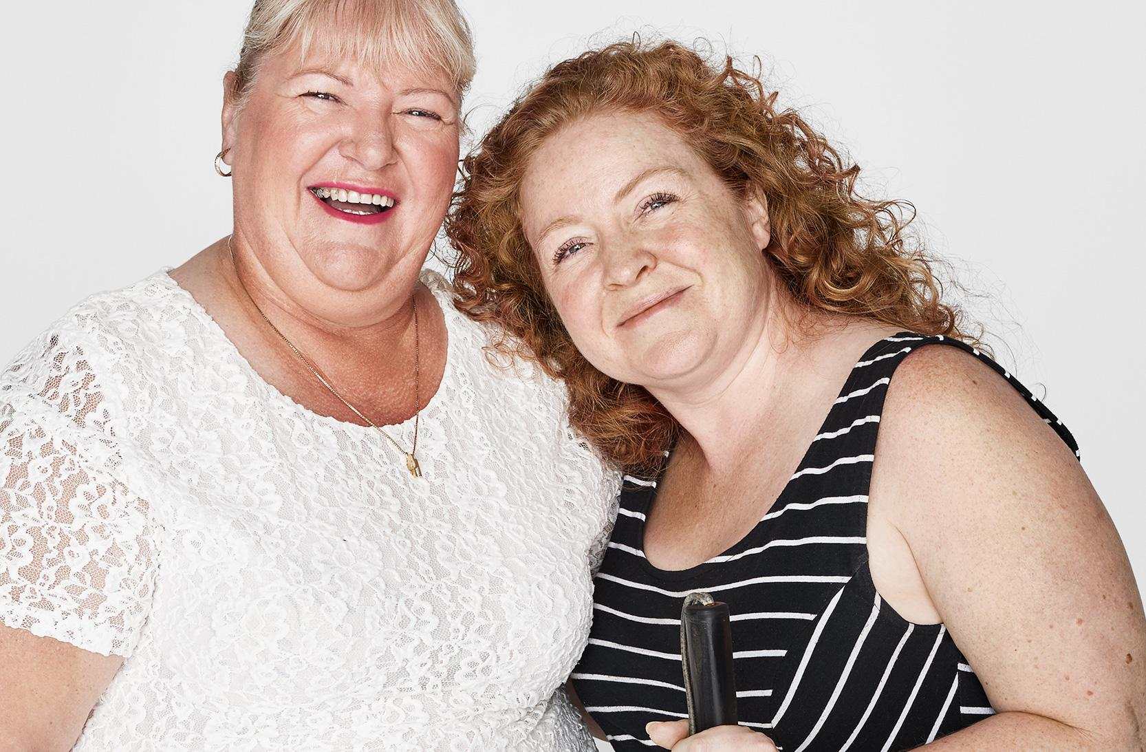 An older woman in a white lace dress and a younger woman in a striped top, holding a white cane, stand close together, smiling.