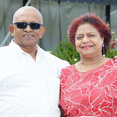 An older couple stands outdoors, smiling. The man wears sunglasses and a white shirt, and the woman wears a red patterned dress.