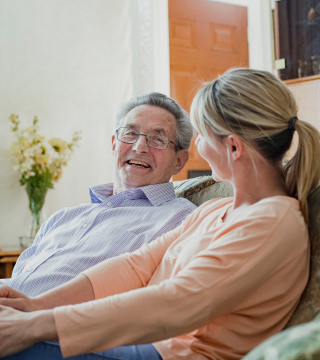A woman is sitting with an older man on the couch. They are facing each other and engaged in discussion.